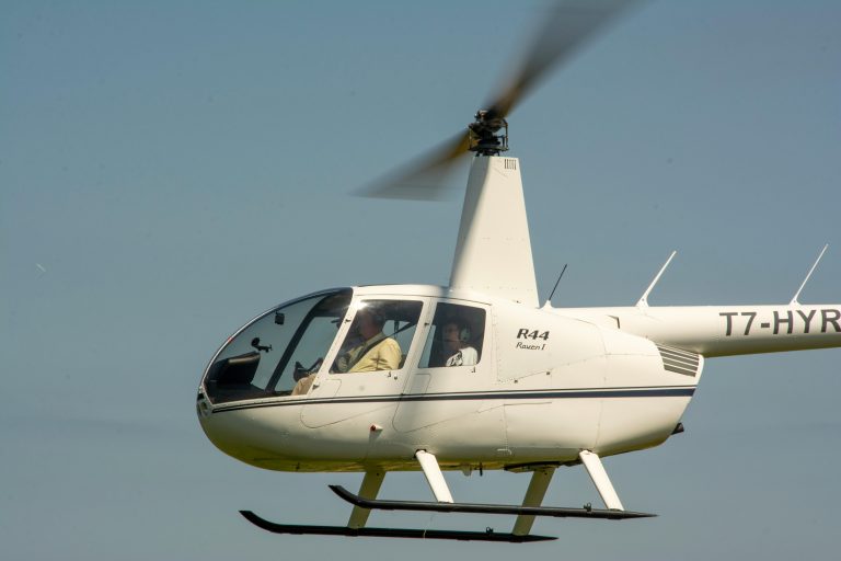 Close-up of a white R44 helicopter in the sky with visible pilots inside.