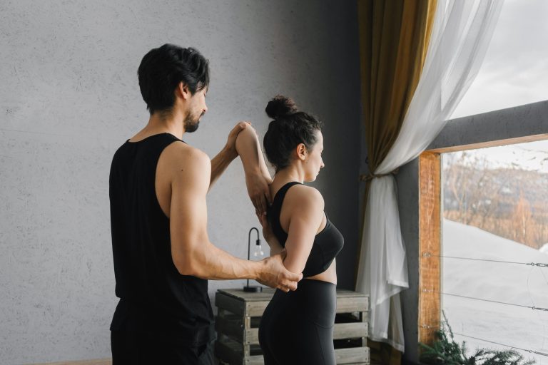 A man and woman practicing partner yoga poses near a large window inside a cozy room.