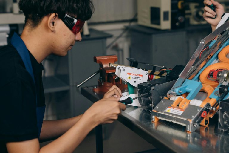 A focused young technician working on electronic equipment in a workshop setting, wearing safety goggles.